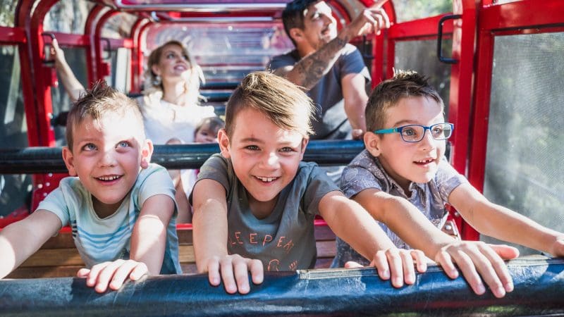 Three happy boys ride a vibrant red attraction at Scenic World, families and Lyrebird Pass holders visible in the lively background.