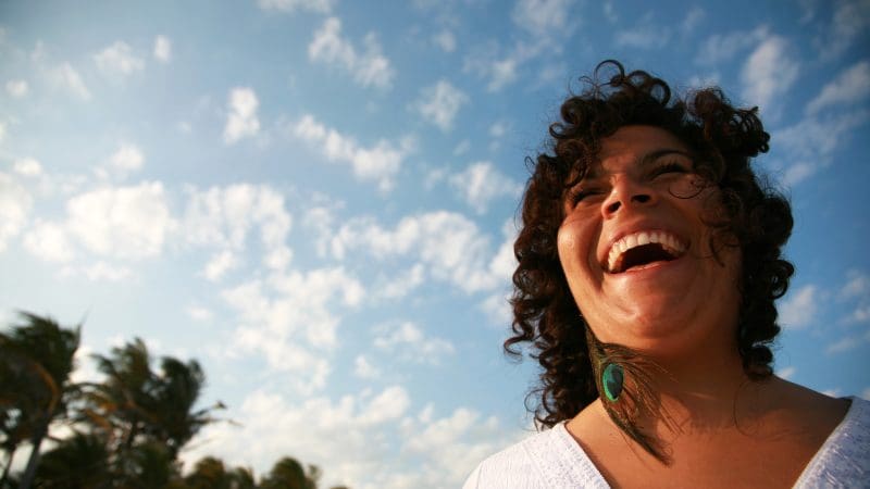 Curly-haired woman laughs joyfully outdoors, enjoying the Hinterland Magic Glow-worm Experience beneath vibrant blue skies.