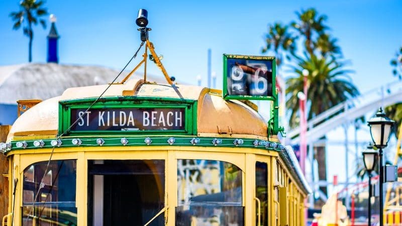 Iconic vintage yellow tram featuring a St Kilda Beach sign, framed by swaying palm trees and clear blue Melbourne skies, epitomising adventure.