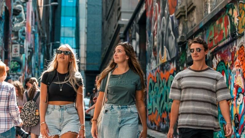 Three fashionable young adults explore a vibrant graffiti alley in Melbourne, wearing trendy summer outfits and sunglasses, soaking up city vibes.