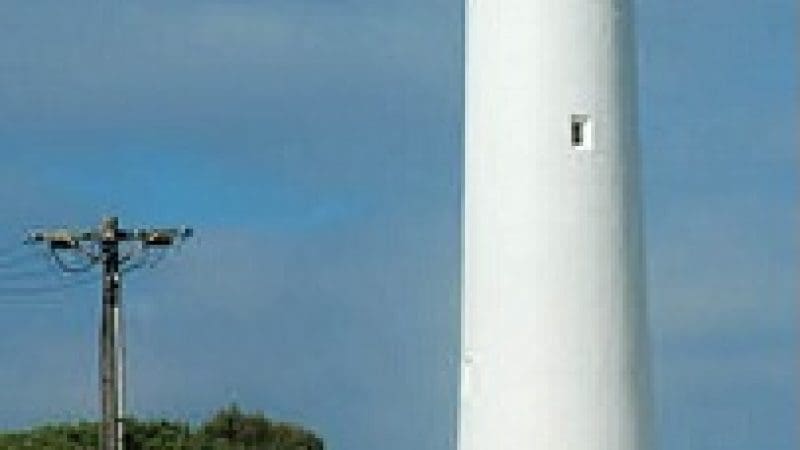 Three tourists stand on a scenic path beside a towering white lighthouse on the Ultimate 2-Day Great Ocean Road Tour in Australia.