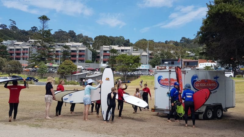 Surfers in wetsuits holding surfboards assemble by the Learn To Surf Tour trailer on a lush, green seaside grassy area.