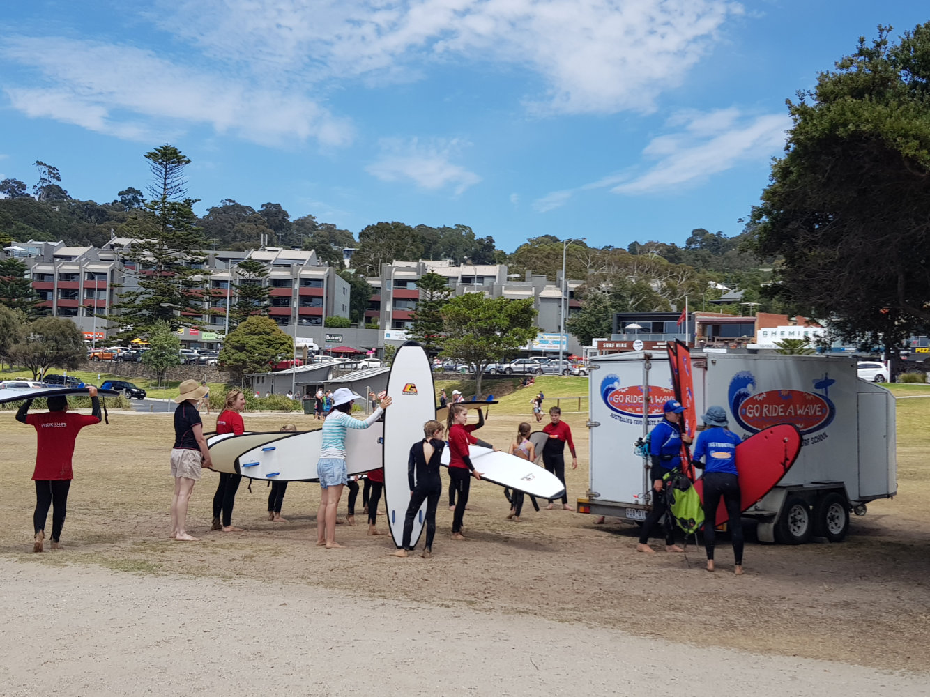 Surf lesson group with surfboards by a Go Ride a Wave trailer in a grassy seaside park during a Learn to Surf Tour.
