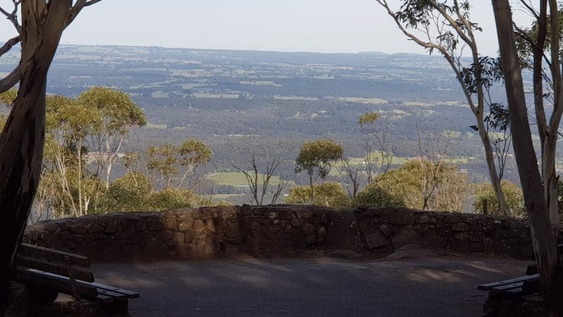Scenic stone lookout with benches amid lush trees, showcasing panoramic Mt Macedon Ranges and Daylesford views under clear skies.