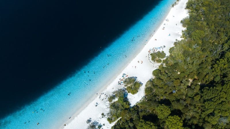 Stunning aerial view of Kgari 5 Day Ultimate Beach with pristine white sand, crystal-clear turquoise water, lush greenery, and visitors enjoying the sun.
