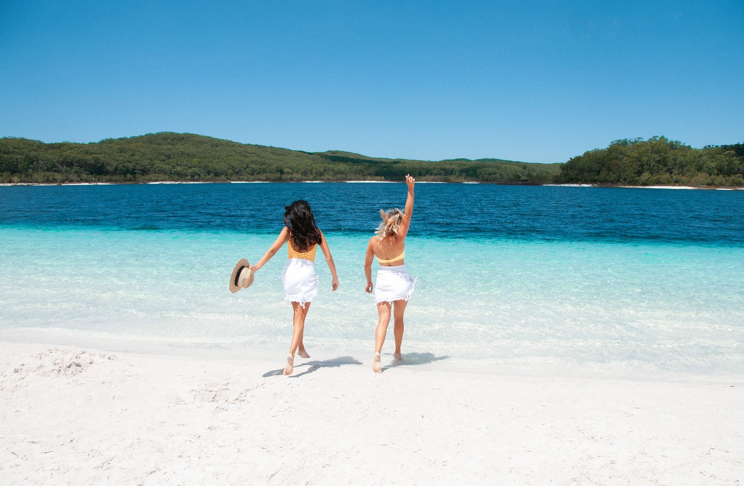 Two women in swimsuits stroll along pristine sand towards crystal-clear blue water on a K'gari Explorer Day Tour departing Rainbow Beach.