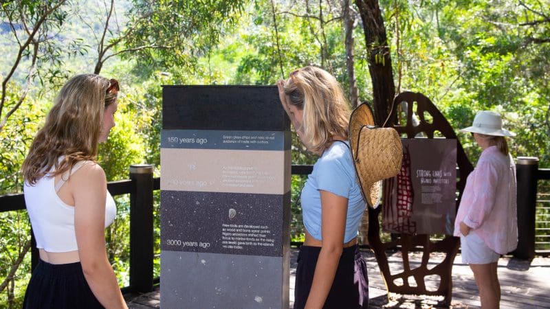 Three women reading informational signs surrounded by lush greenery, sunlight illuminating the Lady Enid Sailing walkway experience.
