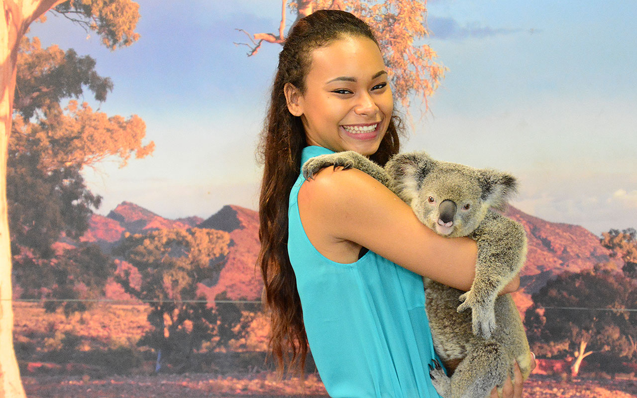 Happy woman in a blue dress holds a koala at Kuranda Koala Gardens, showcasing her 1 Day Kuranda Wildlife Experience Pass.
