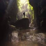 Hiker stands on a rock in Twister and Rocky Creek Canyons, lush ferns and sunlit, crystal-clear water flowing around them.