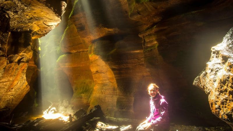 Hiker rests on a sunlit rock in Double Canyon Adventure, vibrant moss-covered walls illuminated by beams in Australia’s Blue Mountains.