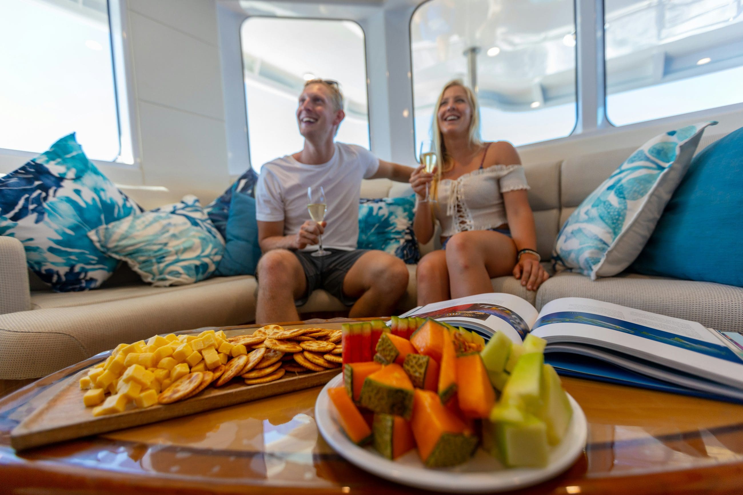 Two guests relax on a luxurious yacht sofa, savouring drinks, gourmet snacks, and a book whilst experiencing the Evolution Gold Class Lounge.