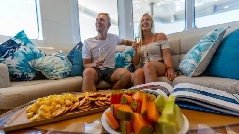 Two guests relax on a luxurious yacht sofa, savouring drinks, gourmet snacks, and a book whilst experiencing the Evolution Gold Class Lounge.