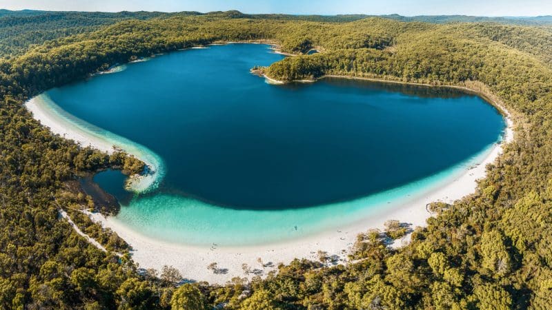 Stunning aerial of Illumina’s heart-shaped blue lake on K'gari 3 Day Getaway, encircled by lush forest and pristine white sands.