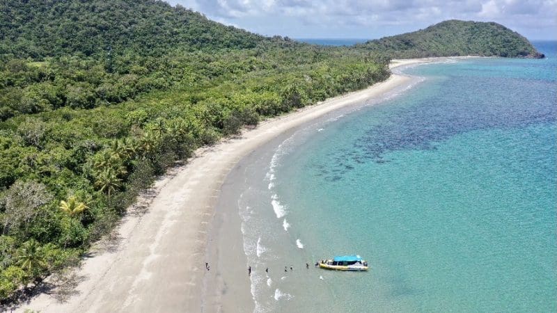 Eco adventure boat anchored by a pristine sandy beach on the Great Barrier Reef, with lush forest and crystal-clear turquoise waters.