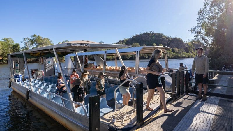 Travellers board a Katherine Gorge cruise riverboat at the dock, surrounded by lush trees and scenic hills on a guided day tour.