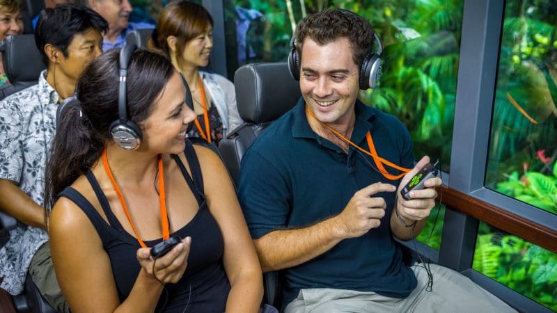 Happy tourists with headphones enjoying a Cape Tribulation Daintree Guided Tour, lush rainforest visible through panoramic coach windows.