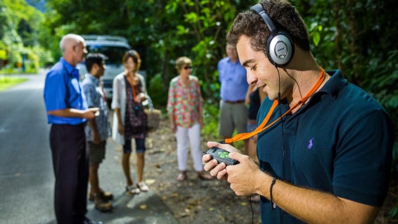 Man with headphones using a device during Cape Tribulation Daintree Guided Day Tour, tour group and lush rainforest scenery in background.