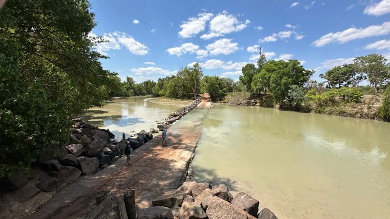 A narrow stone causeway spans a tranquil river beneath clear blue skies, ideal for Kakadu Litchfield Tour camping and nature adventures.