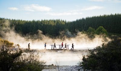 Six people cross a scenic bridge over steaming water; lush trees and greenery surround them beneath a vibrant blue sky.