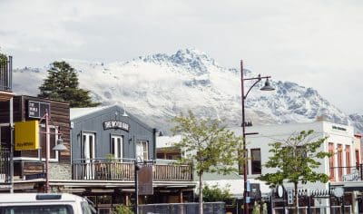 Vibrant city centre street lined with shops and cafés, scenic snowy mountain backdrop, lush trees, and parked cars in the foreground.