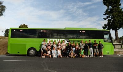 A cheerful group stands together, smiling for a photo in front of a vibrant green coach under clear skies, ready for adventure.