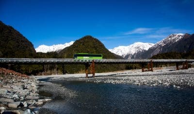 An image of a Kiwi bus crosses a scenic bridge spanning a wide river, with urban skyline and lush greenery visible in the background.