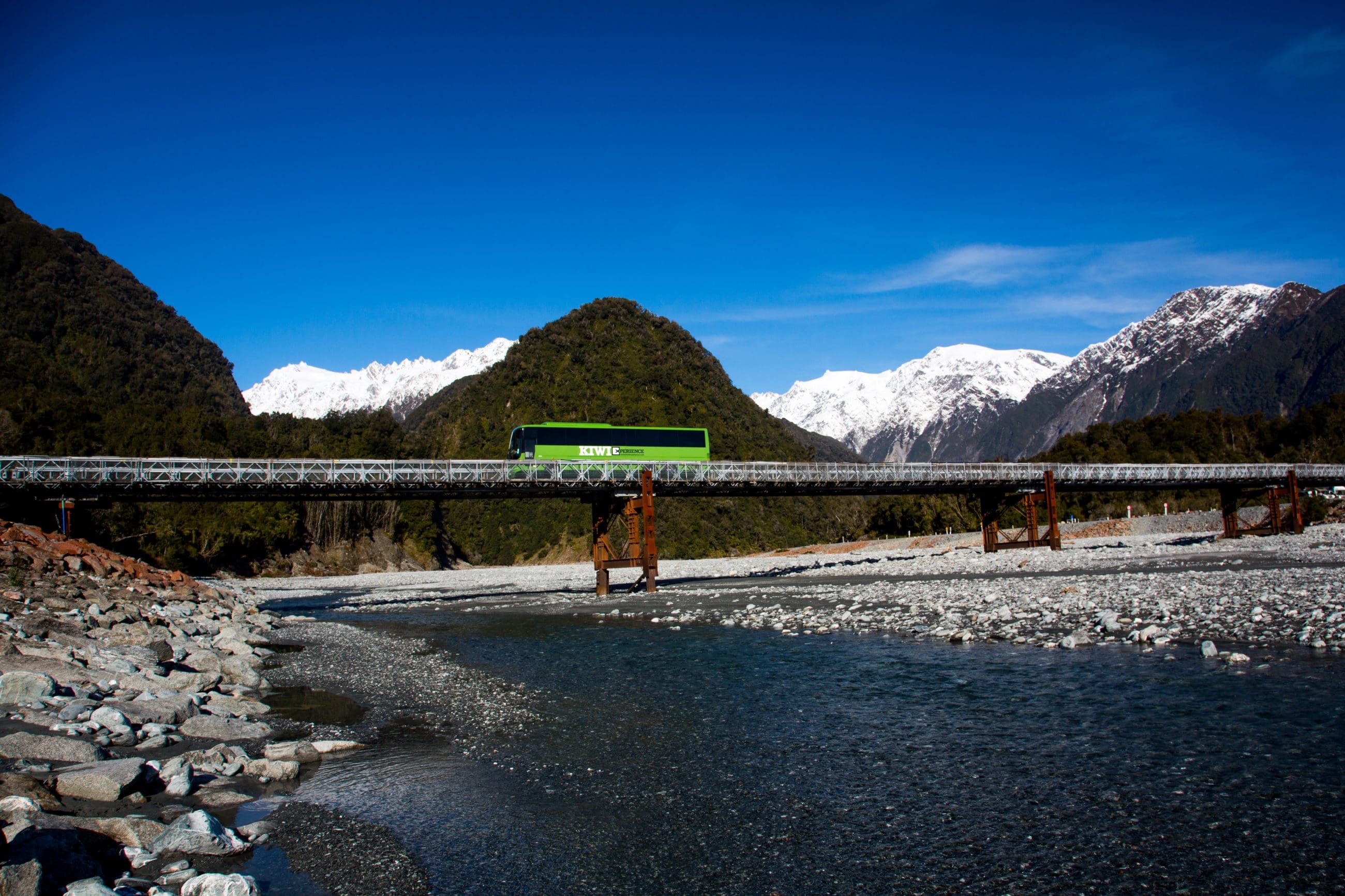 A fully loaded bus travels across a scenic bridge spanning a flowing river, showcasing transport over water in a picturesque setting.