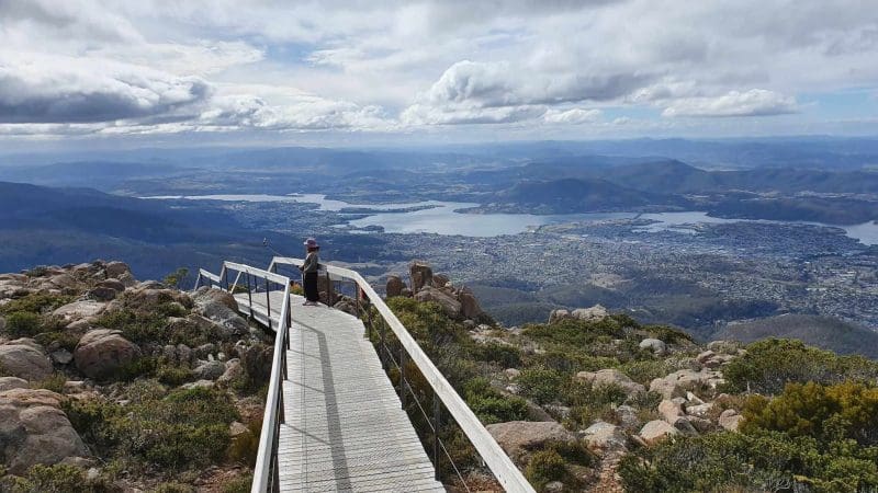 Traveller stands on scenic wooden boardwalk, overlooking stunning mountain ranges and cityscape, reminiscent of Gordon Dam Lake Pedder tour.