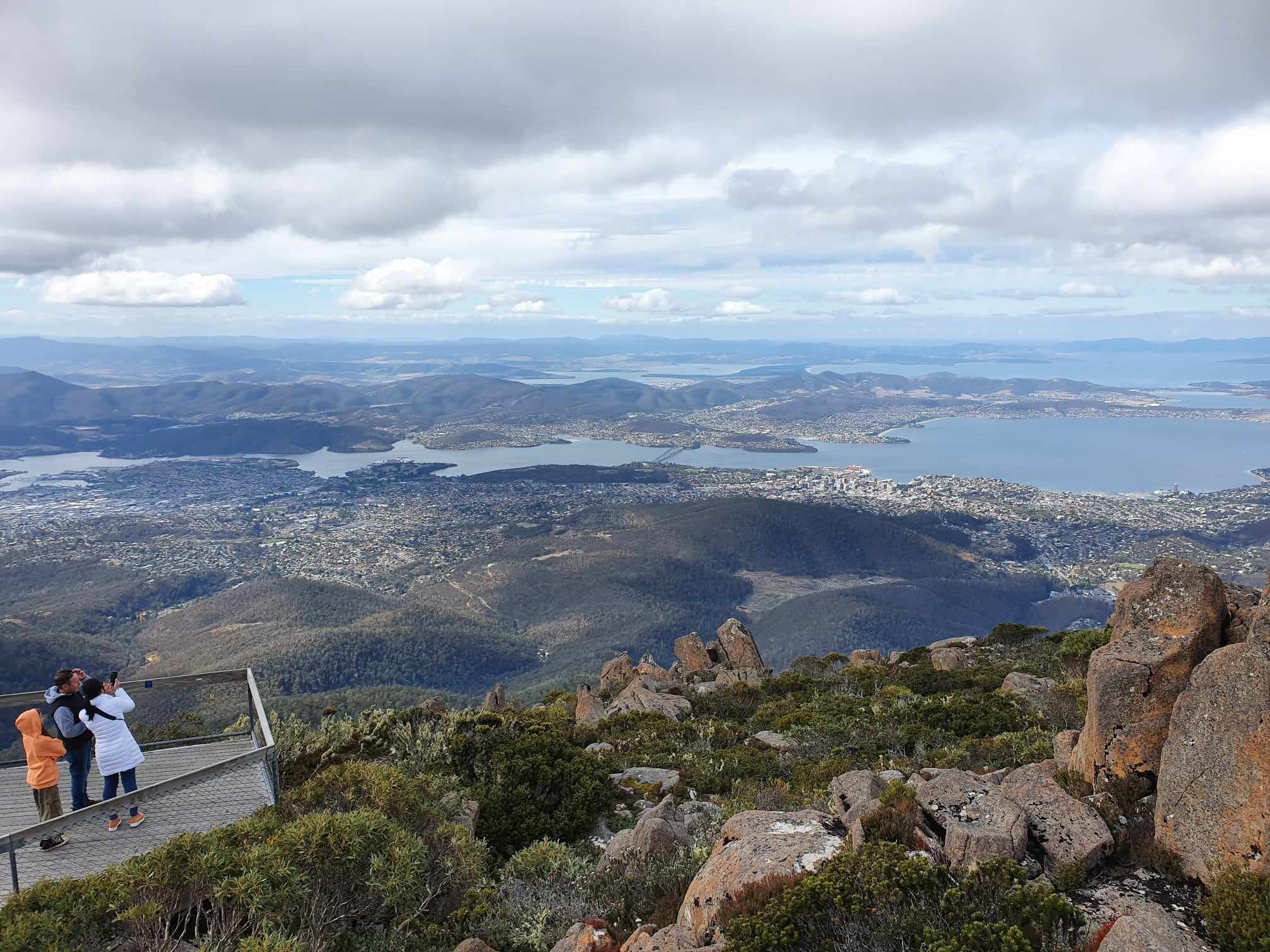 Visitors savour panoramic views during a Mt Wellington Afternoon Driving Tour, overlooking Hobart city, Derwent River, and rolling hills below dramatic clouds.