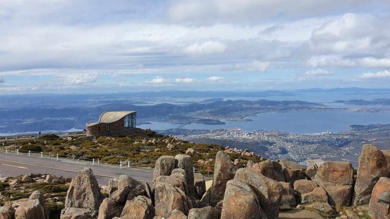 Breathtaking rocky mountain summit with a scenic hut, panoramic views of Lake Pedder on a guided wilderness day tour beneath cloudy skies.