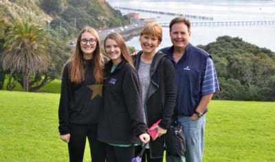 Four people smiling on lush green grass with the iconic Auckland city skyline, waterfront, and trees in the scenic background.