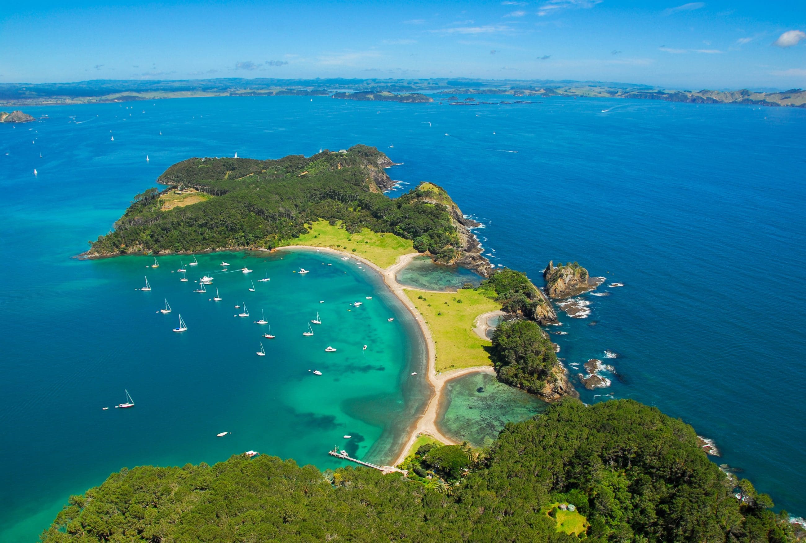 Stunning aerial view of Bay of Islands, featuring vibrant lush greenery, sailboats on crystal-clear turquoise waters, and pristine sandy beach.