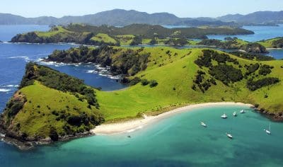 Stunning aerial view of a lush green island in New Zealand’s Bay of Islands, sandy beaches, turquoise waters, and anchored boats.