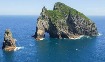 Iconic rocky island with natural sea arch in New Zealand’s Bay of Islands, vibrant greenery, crystal-blue ocean, boats, and clear skies.