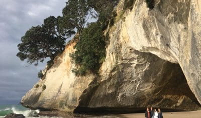 Group of four tourists walking along Coromandel beach near iconic coastal rock archway and lush trees, enjoying scenic New Zealand views.