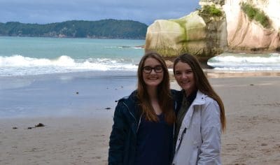 Two happy young women posing on Coromandel beach, golden sand, iconic rock formation, scenic ocean backdrop, New Zealand travel.