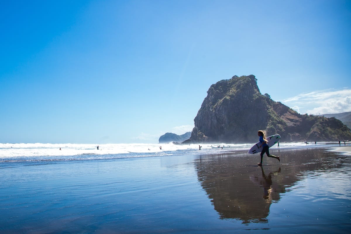 Surfer with board walks along the wet, scenic West Coast beach near crashing waves and a rocky hill beneath clear blue sky.