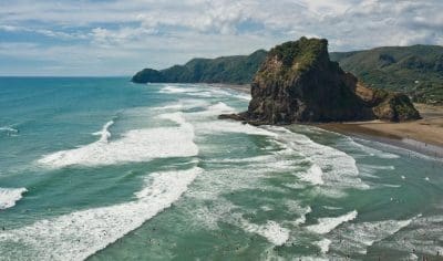 Dramatic waves crash onto a sandy West Coast beach as people swim near a towering rocky hill under a partly cloudy coastal sky.