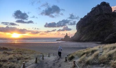 A person in a hoodie strolls along a sandy West Coast path towards the beach at sunset, showcasing stunning coastal scenery.
