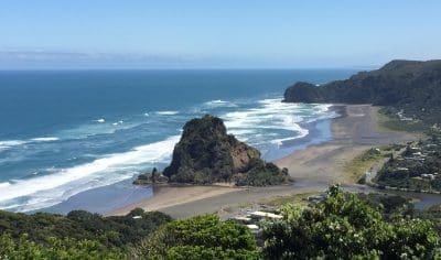 Breathtaking West Coast beach with rocky outcrop, rolling waves, lush greenery, and scenic hills beneath a vibrant blue sky.