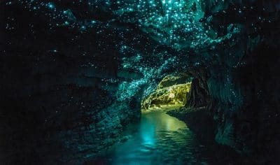 Waitomo-like dark cave with vivid blue-green bioluminescent glow-worms illuminating walls and stream, creating a magical reflection.