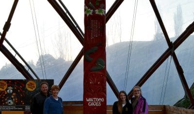 Four visitors pose beside a striking red totem pole inside the Kiwi Bird House’s wooden dome with panoramic glass walls.