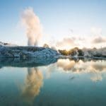 Rotorua geothermal hot spring at sunrise, steam rising and mirrored on tranquil waters, framed by lush greenery and rugged rocks.