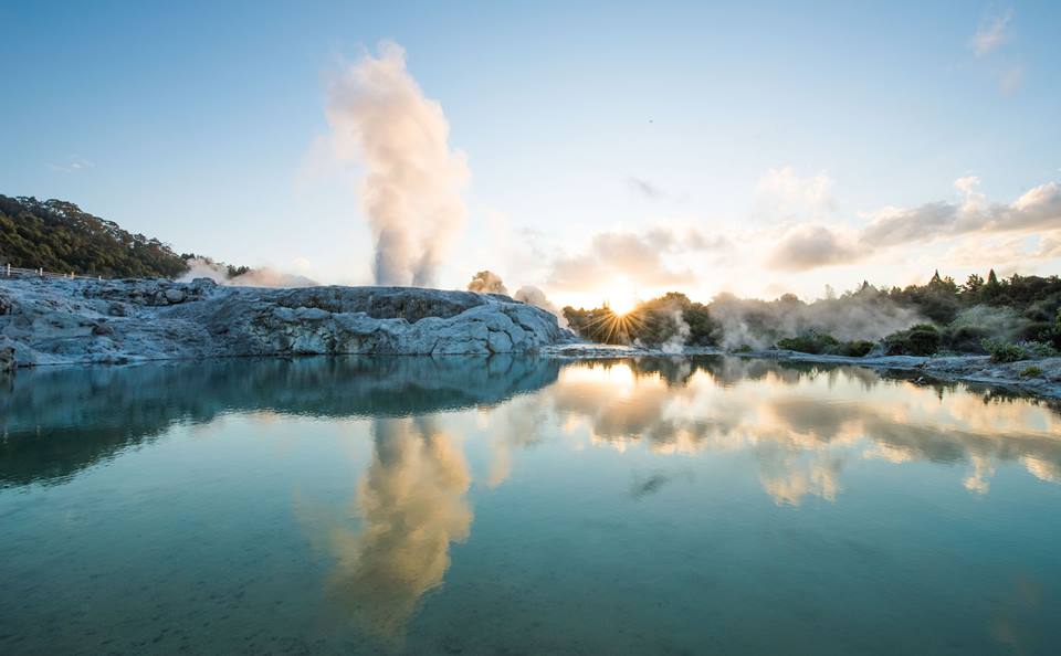 Rotorua geothermal hot spring at sunrise, steam rising and mirrored on tranquil waters, framed by lush greenery and rugged rocks.