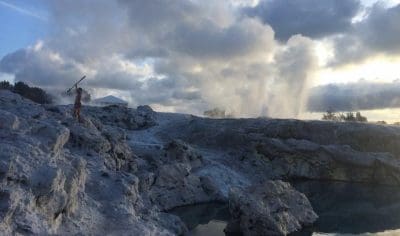 Explorer on rugged rocks beside steaming Rotorua geothermal pools, dramatic sunset clouds overhead; top New Zealand travel destination.