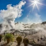 Rotorua geothermal hot springs emit steam under a sunny sky, surrounded by lush greenery and fences, showcasing New Zealand’s beauty.