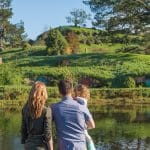 A family enjoys the scenic Hobbiton Movie Set with iconic hillside hobbit houses by a tranquil pond under a clear blue sky in New Zealand.