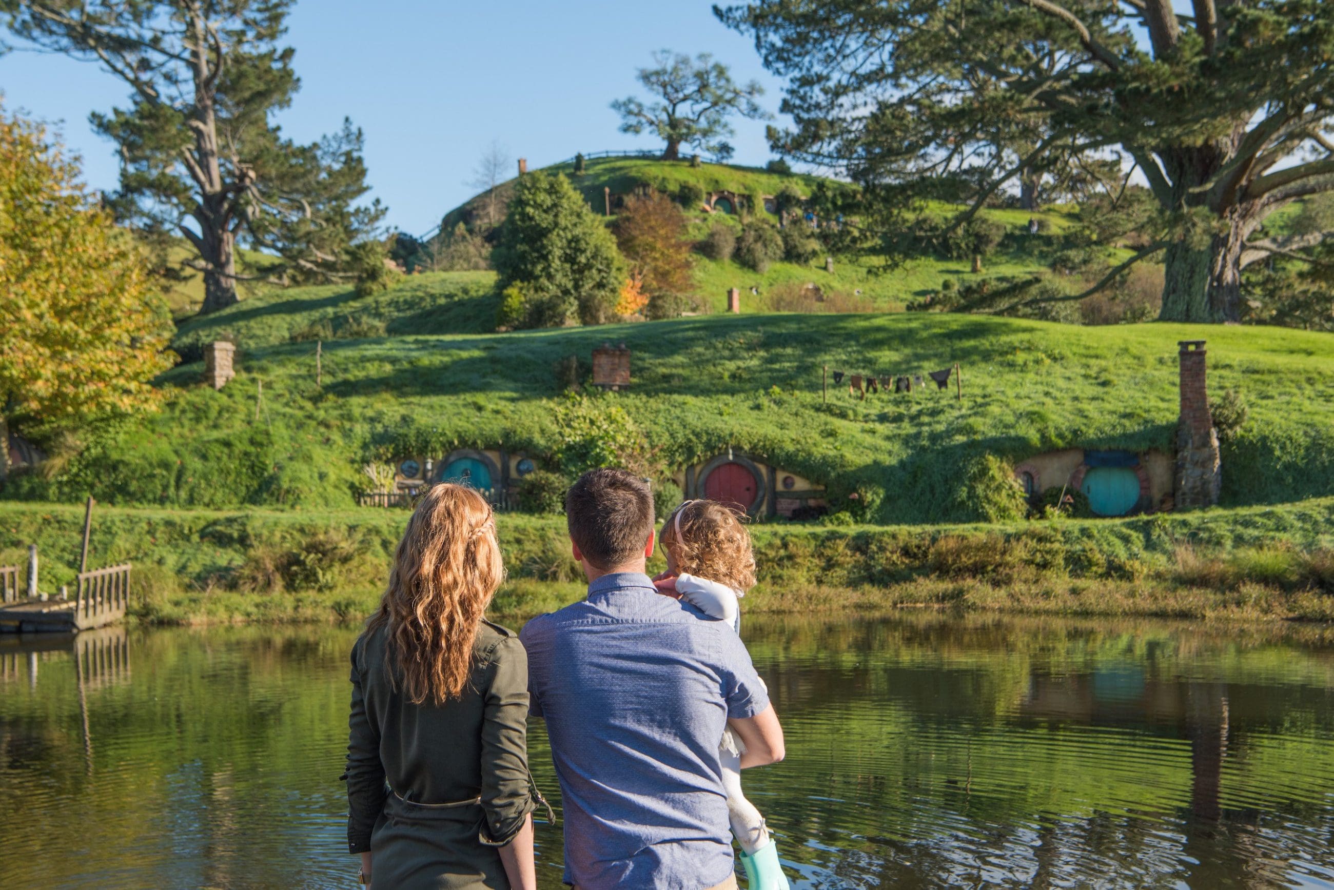 A family enjoys the scenic Hobbiton Movie Set with iconic hillside hobbit houses by a tranquil pond under a clear blue sky in New Zealand.