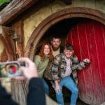 Three happy visitors pose by a red Hobbit door at the Hobbiton Movie Set as their photo is captured on a smartphone for memories.