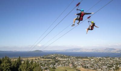 Adventurers zipwire high above Rotorua's stunning lake and lush hills, experiencing thrilling outdoor activities under a clear blue sky.